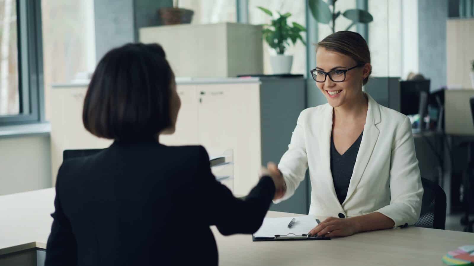 Zwei Frauen, die sich über einen Schreibtisch hinweg die Hand geben